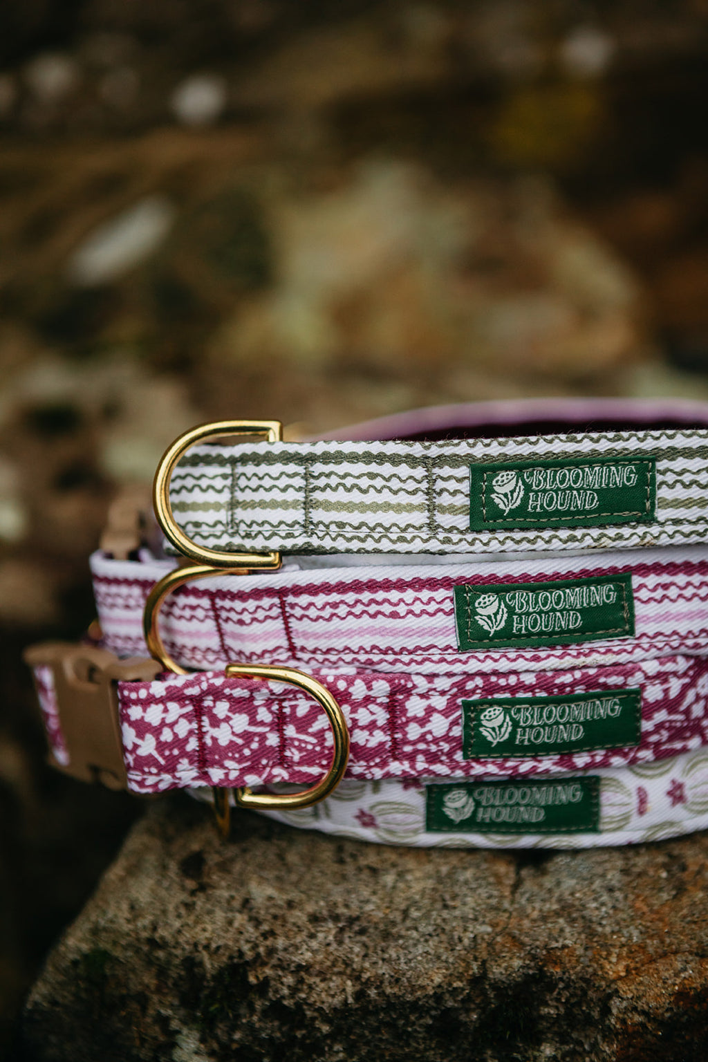 Multiple dog collars on a rock with Blooming Hound label, brass hardware, and acetal buckle. Patterns are Moss Trail, Heather Trail, Seed Pod and Heather.
